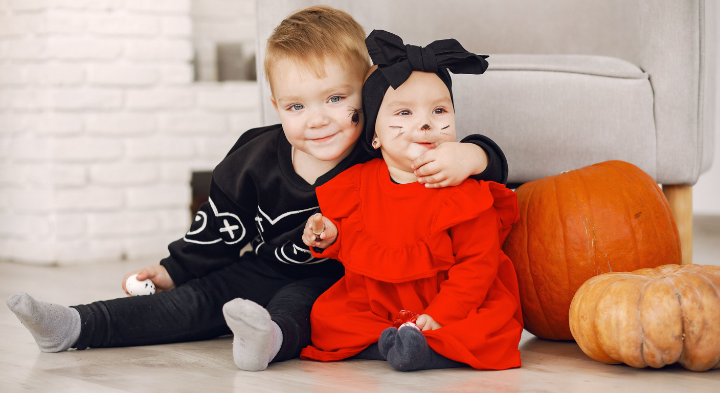 Two toddlers in Halloween costumes with painted faces sitting on the floor beside large orange pumpkins.