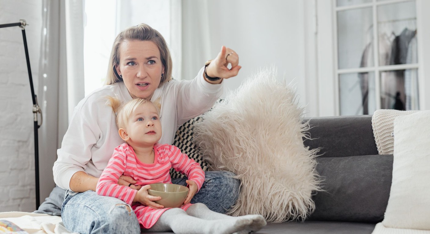 A mother points at the TV and narrates while her toddler watches from her lap