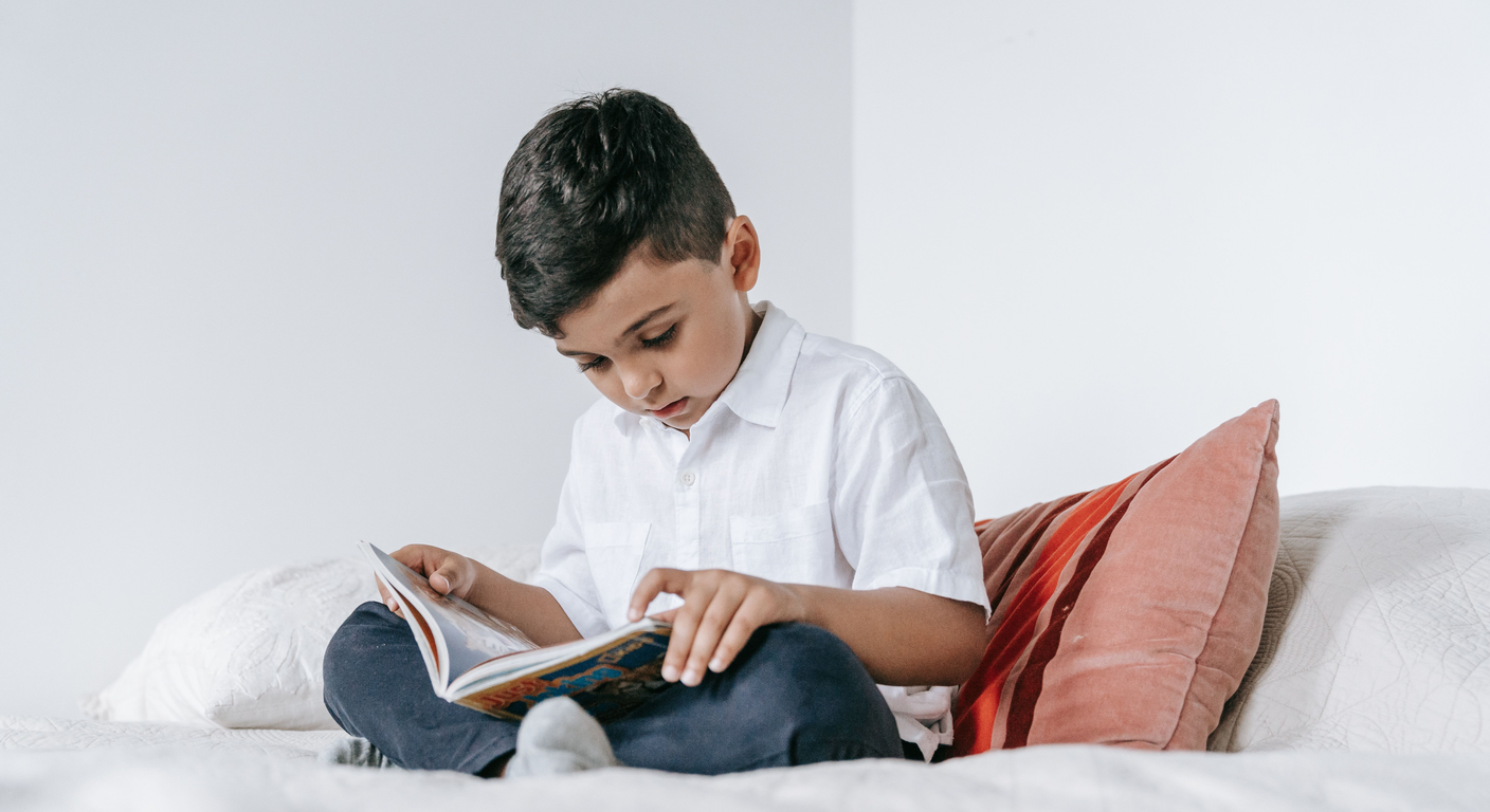 A school-age boy sitting cross-legged on a bed, concentrating on an open book.