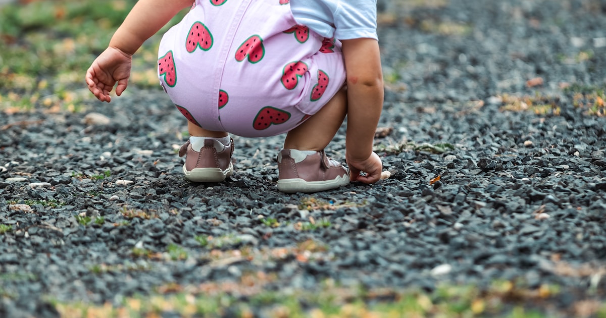 A baby in a strawberry romper crouches on gravel.